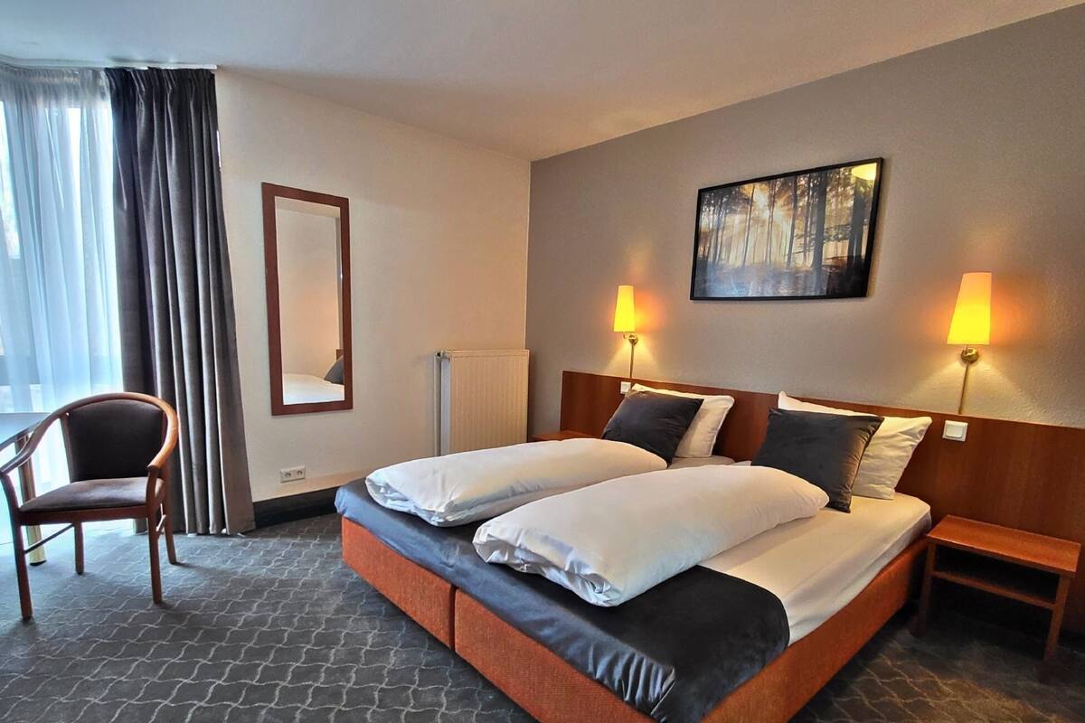 A hotel room with a double bed, two bedside lamps, a chair, a large mirror, and a painting on the wall.