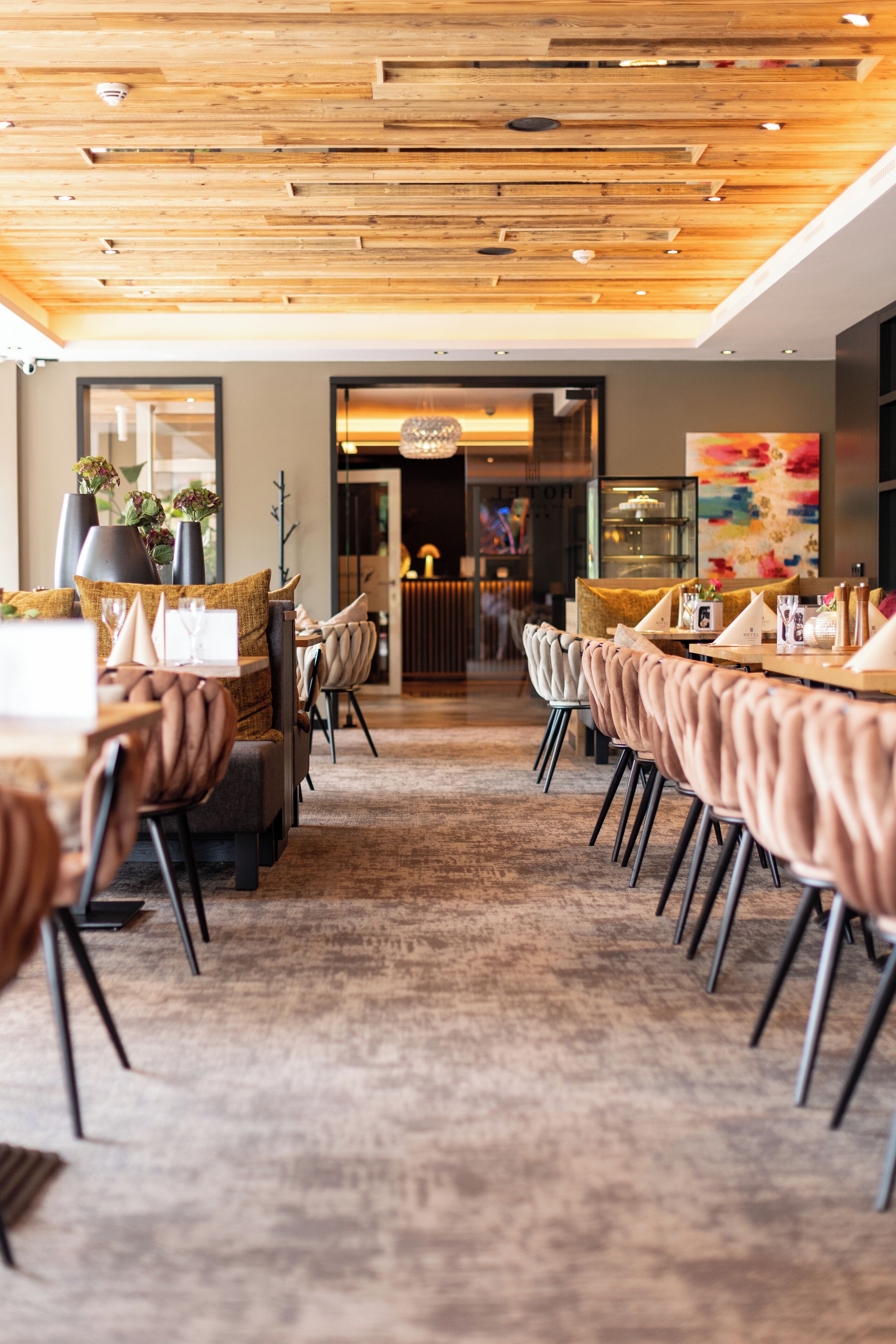 Een modern restaurant met houten plafond, lange tafels en stoelen met kussens.