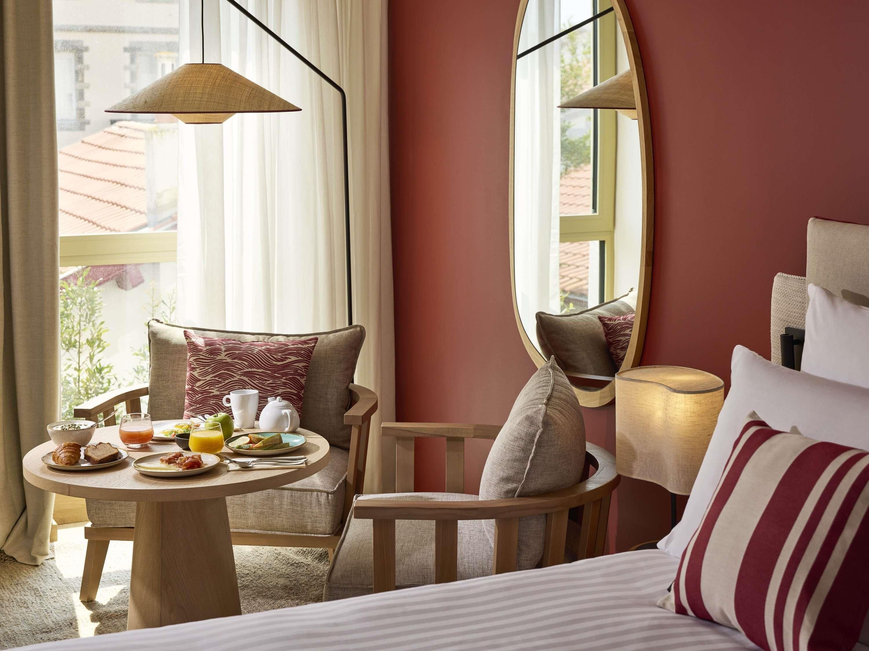 A hotel room with a bed, a round table with breakfast, a chair, a lamp, and a window with curtains.