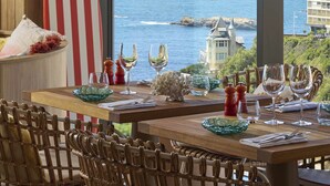 A dining area with a view of the sea, a surfboard, and a table set with glasses and cutlery. - Le Talaia Hôtel & Spa Biarritz - MGallery Collection (Biarritz)