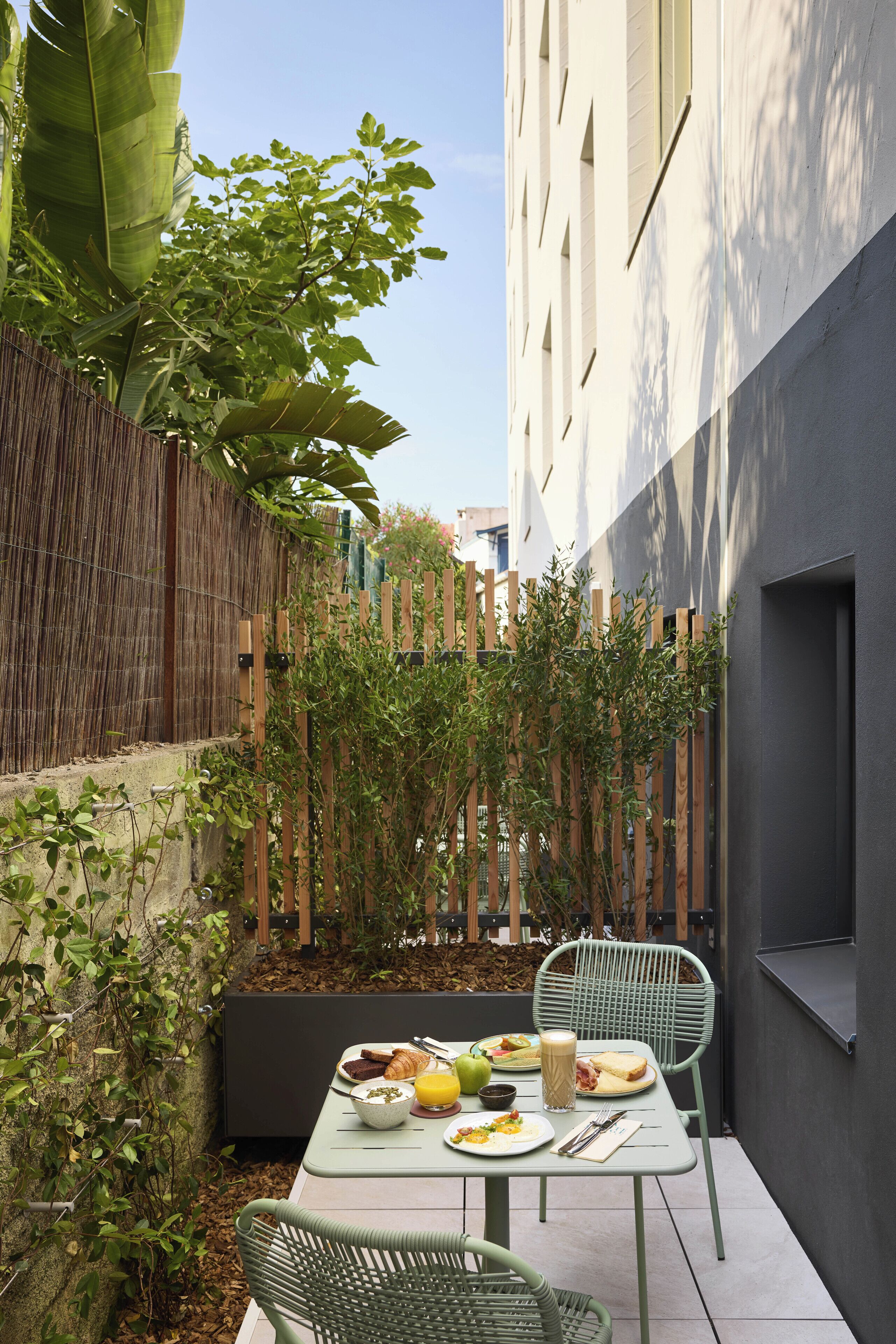 Un petit espace extérieur aménagé pour prendre les repas, avec une table pour deux, entouré d’une clôture en bois et de verdure.