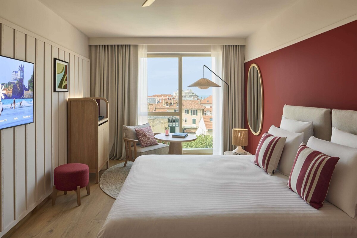 A hotel room with a bed, a TV, a desk, a chair, a round table, and a view of buildings through the window.