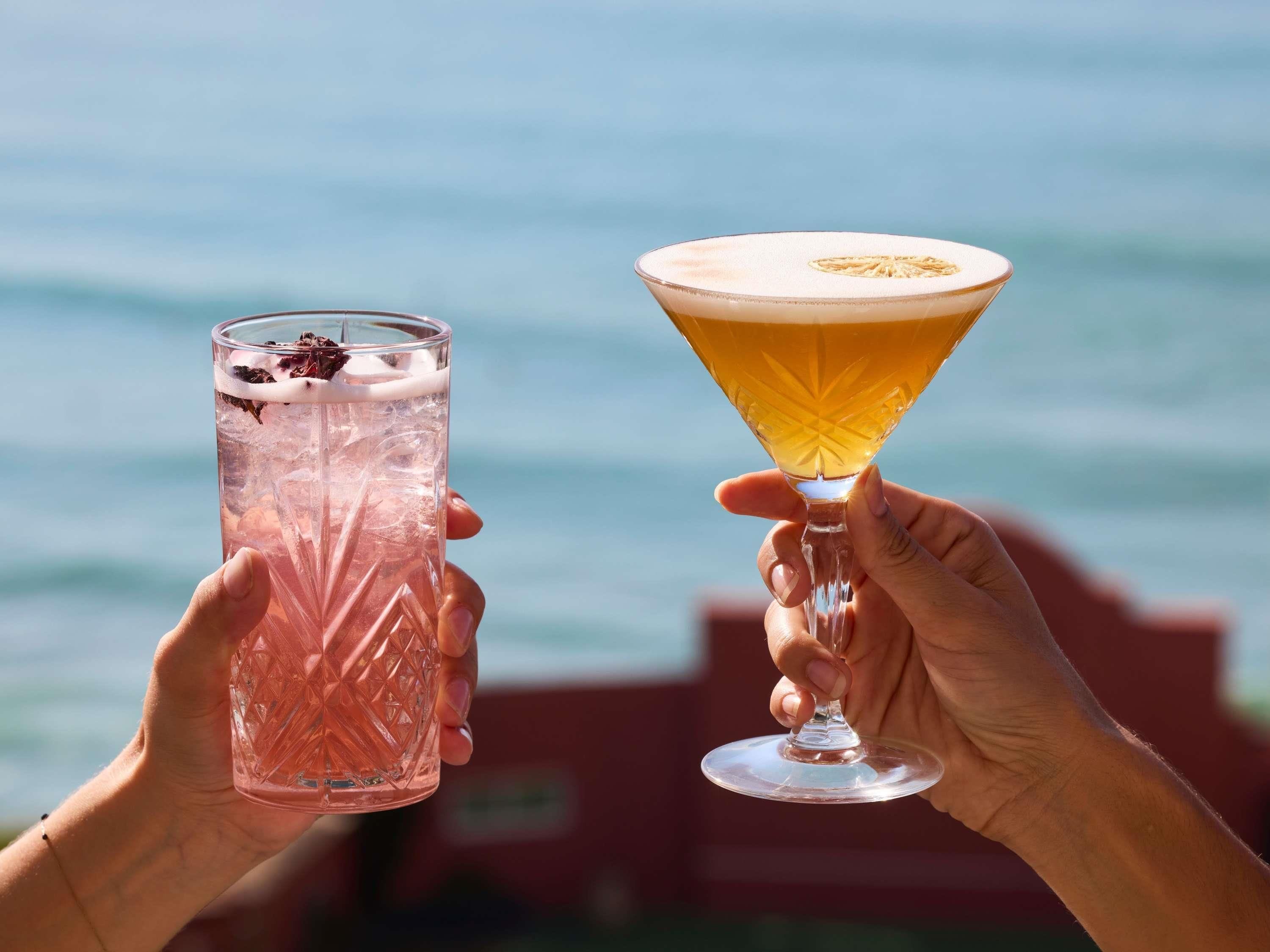 Due mani che sorreggono un bicchiere alto e testurizzato e un bicchiere da martini con un liquido dorato, con sullo sfondo uno specchio d'acqua.