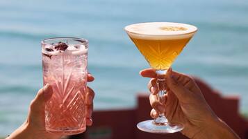 Due mani che sorreggono un bicchiere alto e testurizzato e un bicchiere da martini con un liquido dorato, con sullo sfondo uno specchio d'acqua.