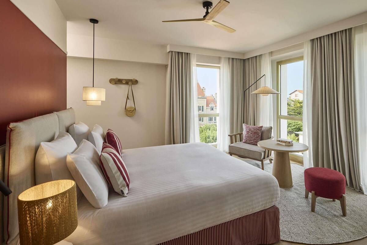 Une chambre d’hôtel avec un grand lit, un bureau, une chaise, un petit tabouret, une lampe, un ventilateur de plafond et une vue sur les bâtiments par les fenêtres.