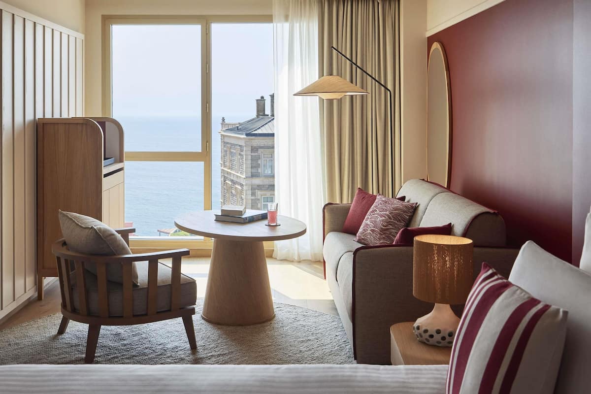 Une chambre d’hôtel avec vue sur la mer, une table ronde avec des livres, un canapé avec des coussins et une lampe.