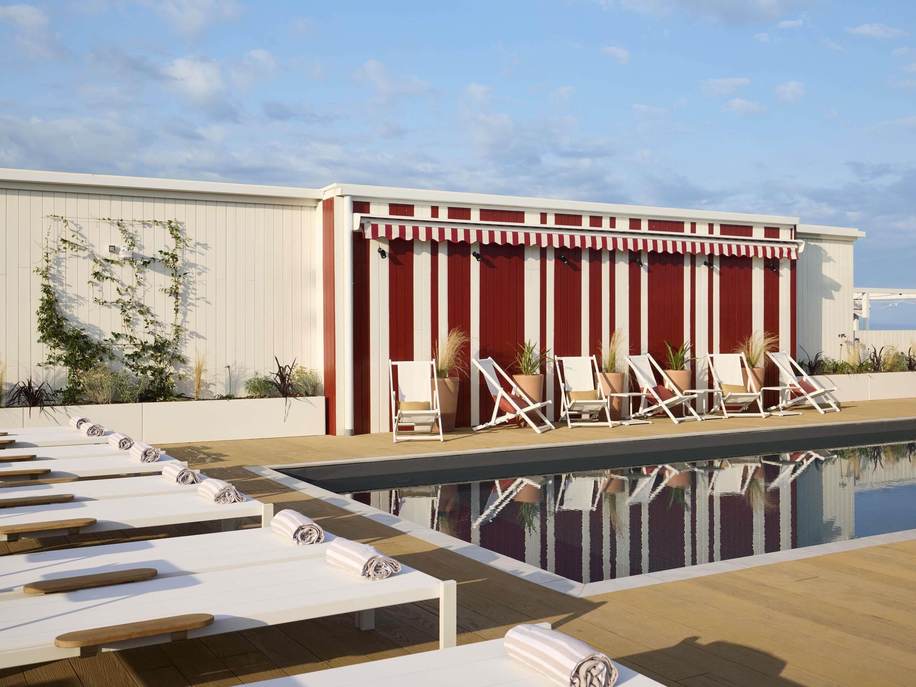 Una piscina en la azotea con sillones de descanso y un edificio de rayas rojas al fondo.