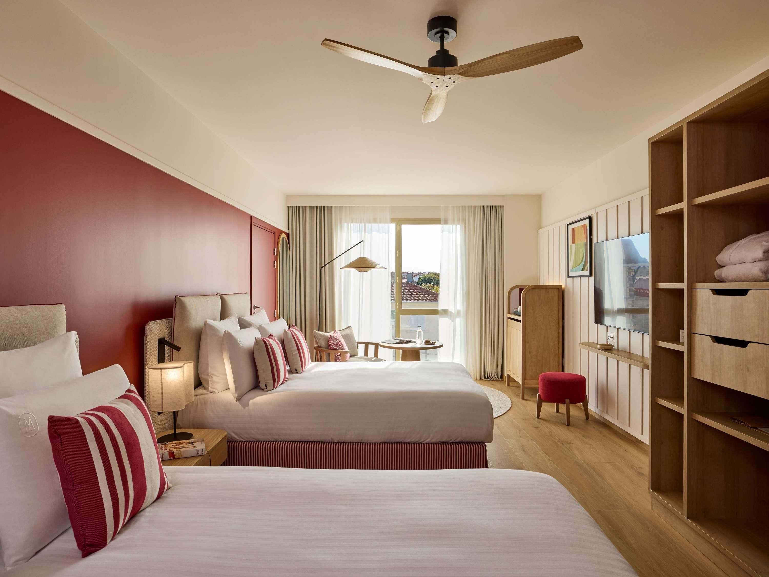 Ein Hotelzimmer mit zwei Betten, einem Fernseher, einem Deckenventilator und einem großen Fenster mit Vorhängen.