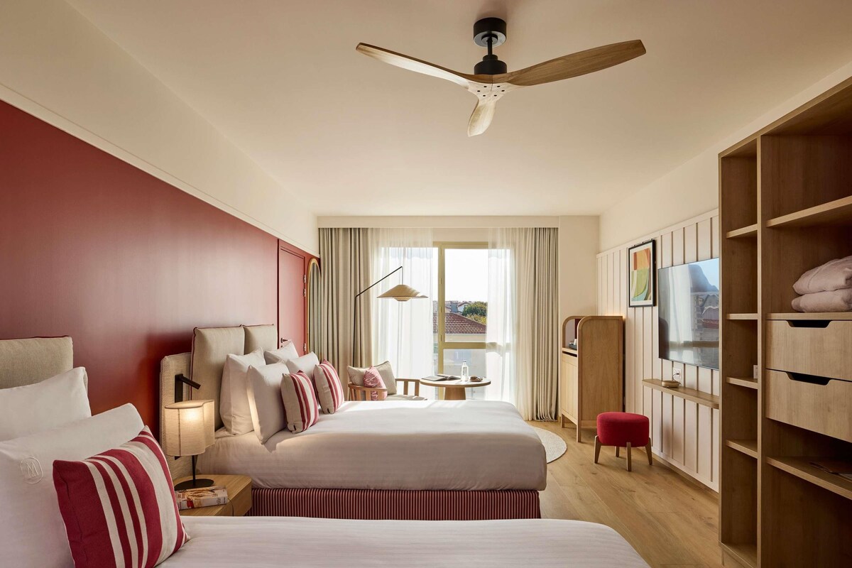 A hotel room with two beds, a TV, a ceiling fan, and a large window with curtains.