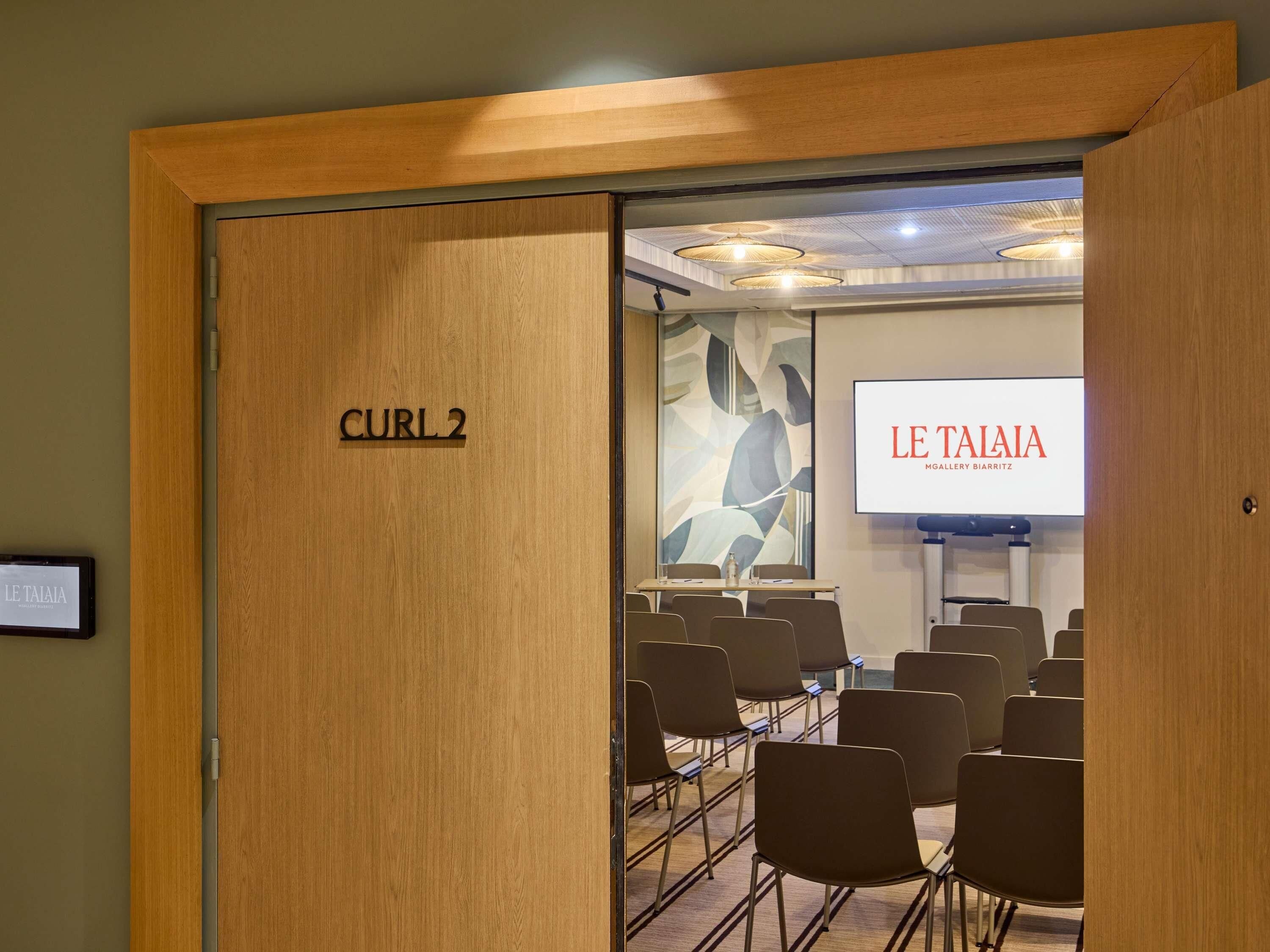 A conference room with a large screen displaying 'LE TAIAIA,' a wooden door labeled 'CURL 2,' and several chairs arranged for meetings.