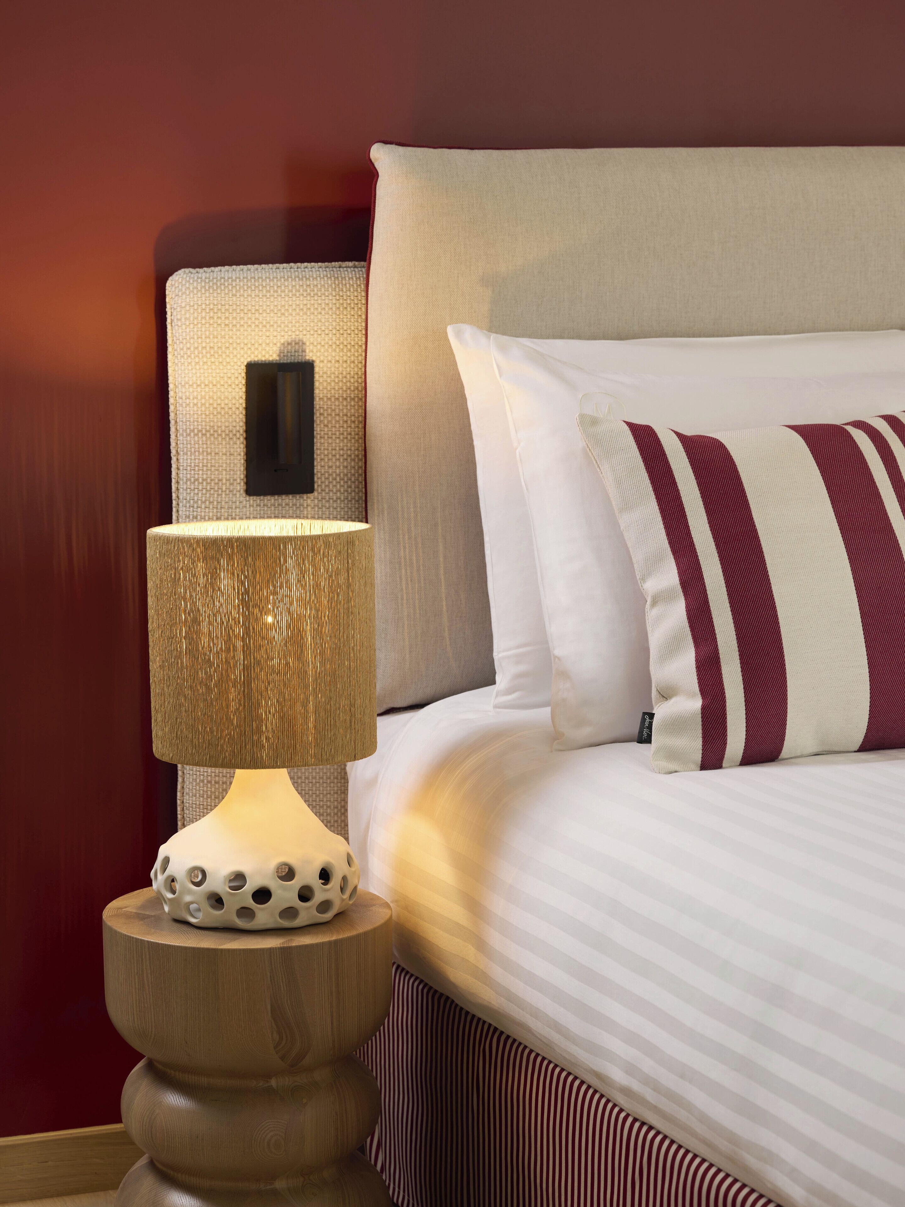 Una cama bien hecha con ropa de cama a rayas y una almohada decorativa, una lámpara de noche con pantalla texturizada y un interruptor de luz en la pared.