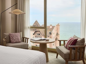 Habitación de hotel con un ventanal grande, una cama, dos sillones, una mesita redonda y una lámpara de pie.
