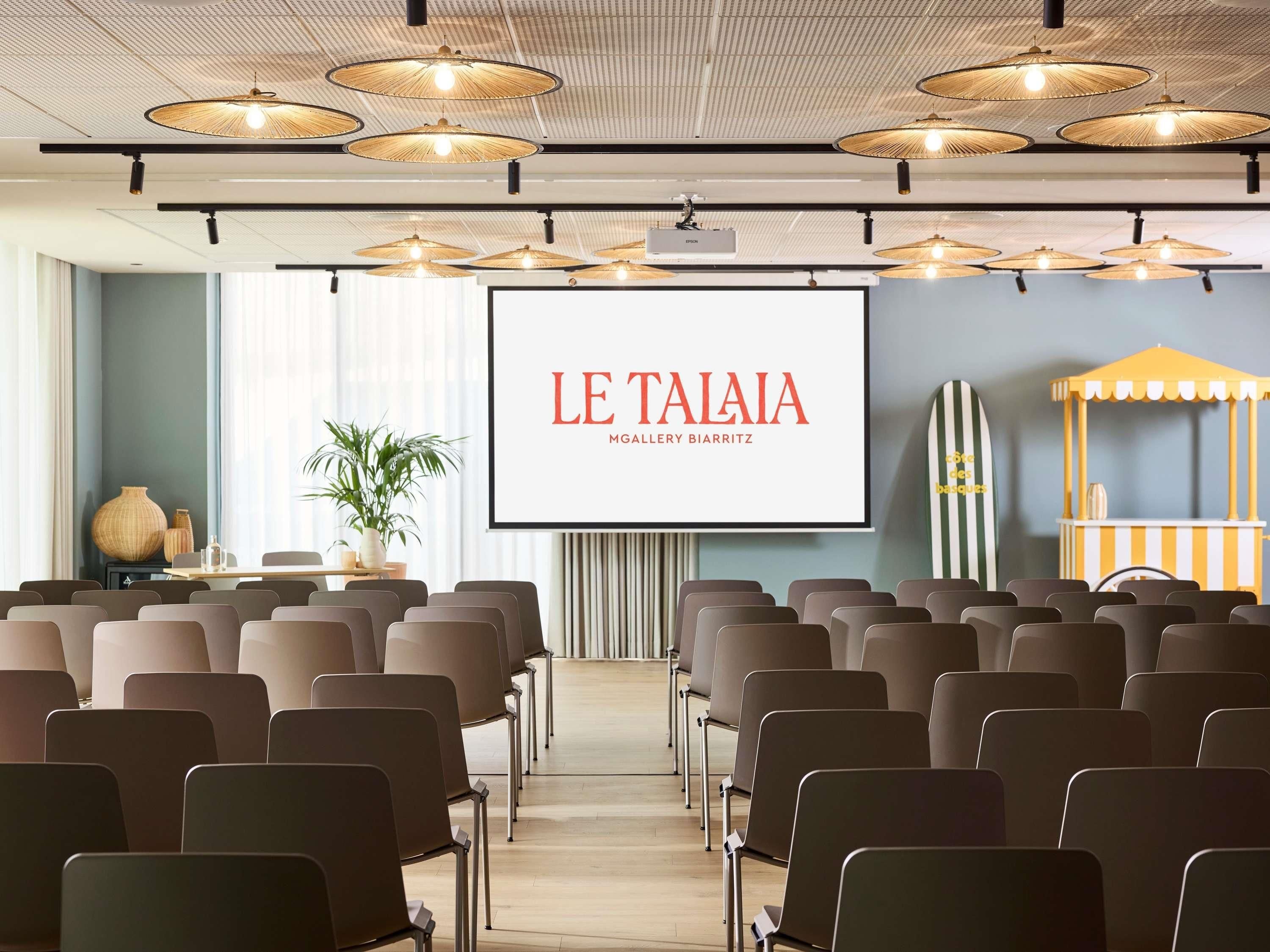 Un salón de conferencias con filas de sillas, una pantalla de proyector mostrando 'LE TAIAIA MGallery BIARRITZ' y una exhibición de tablas de surf.