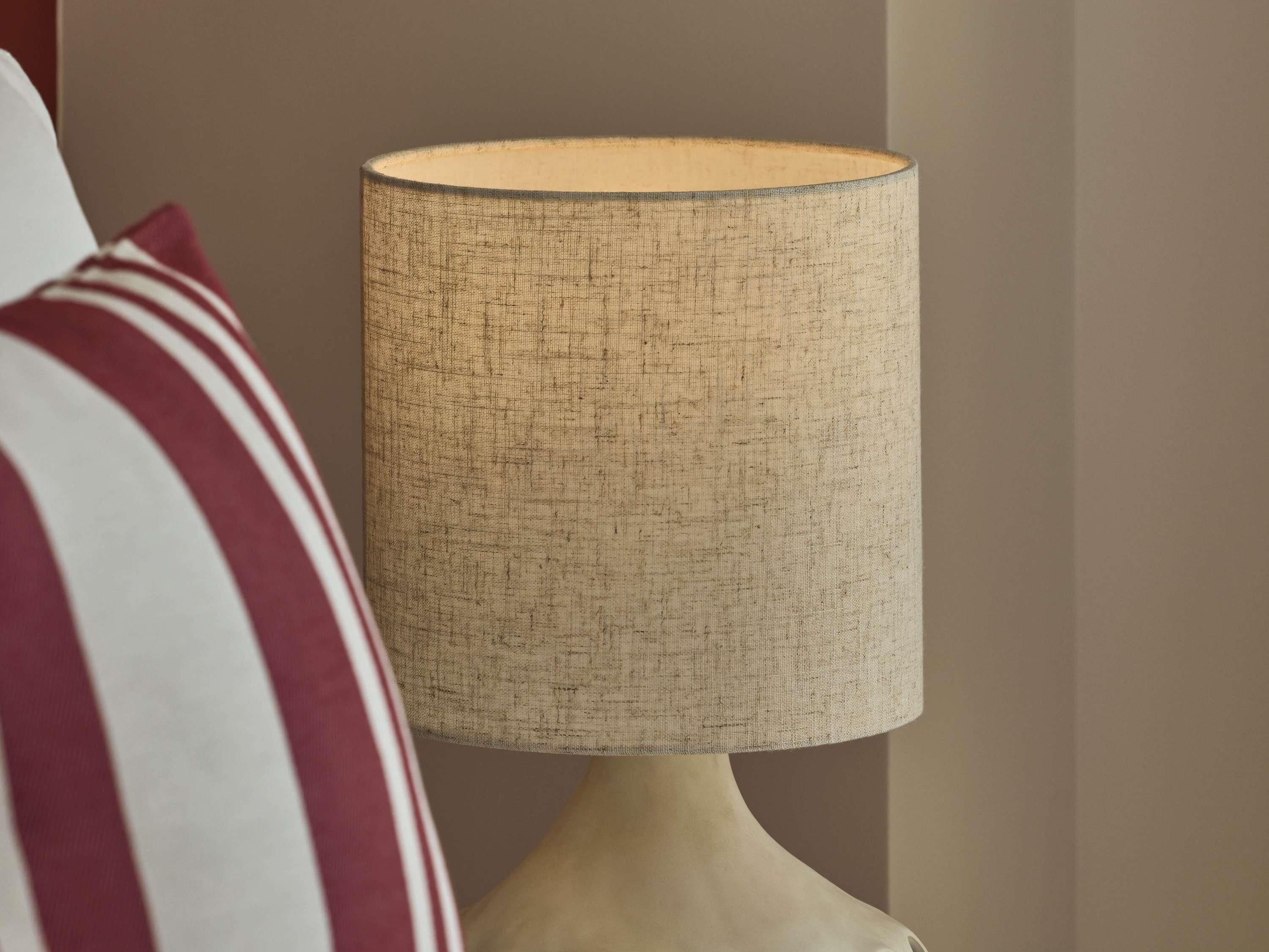 Uma luminária com cúpula de tecido texturizado, base branca e almofada listrada em primeiro plano.