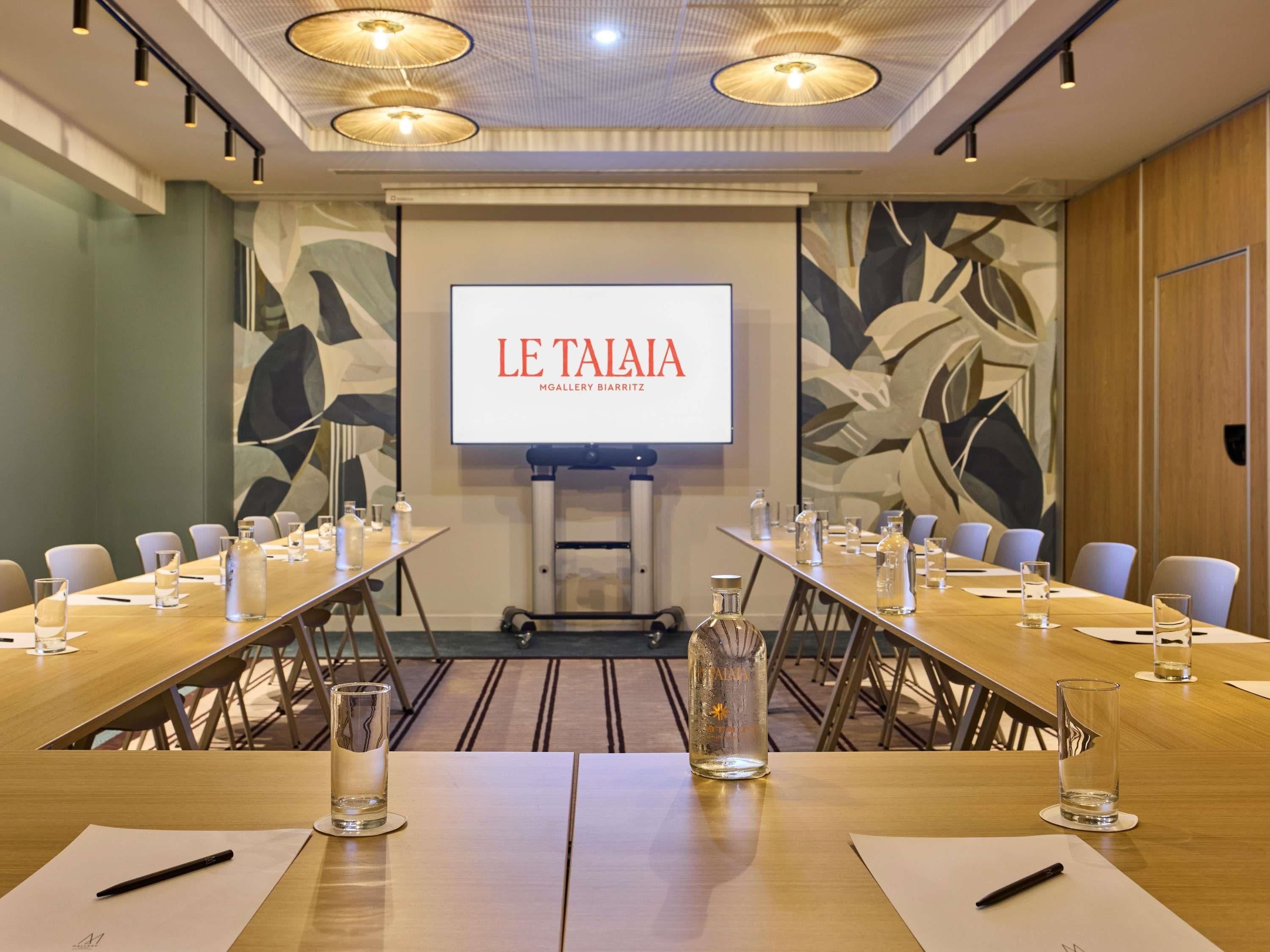 Un salón de conferencias con una pantalla grande que muestra 'LE TAIA', una mesa larga con sillas y un carrito de proyector.