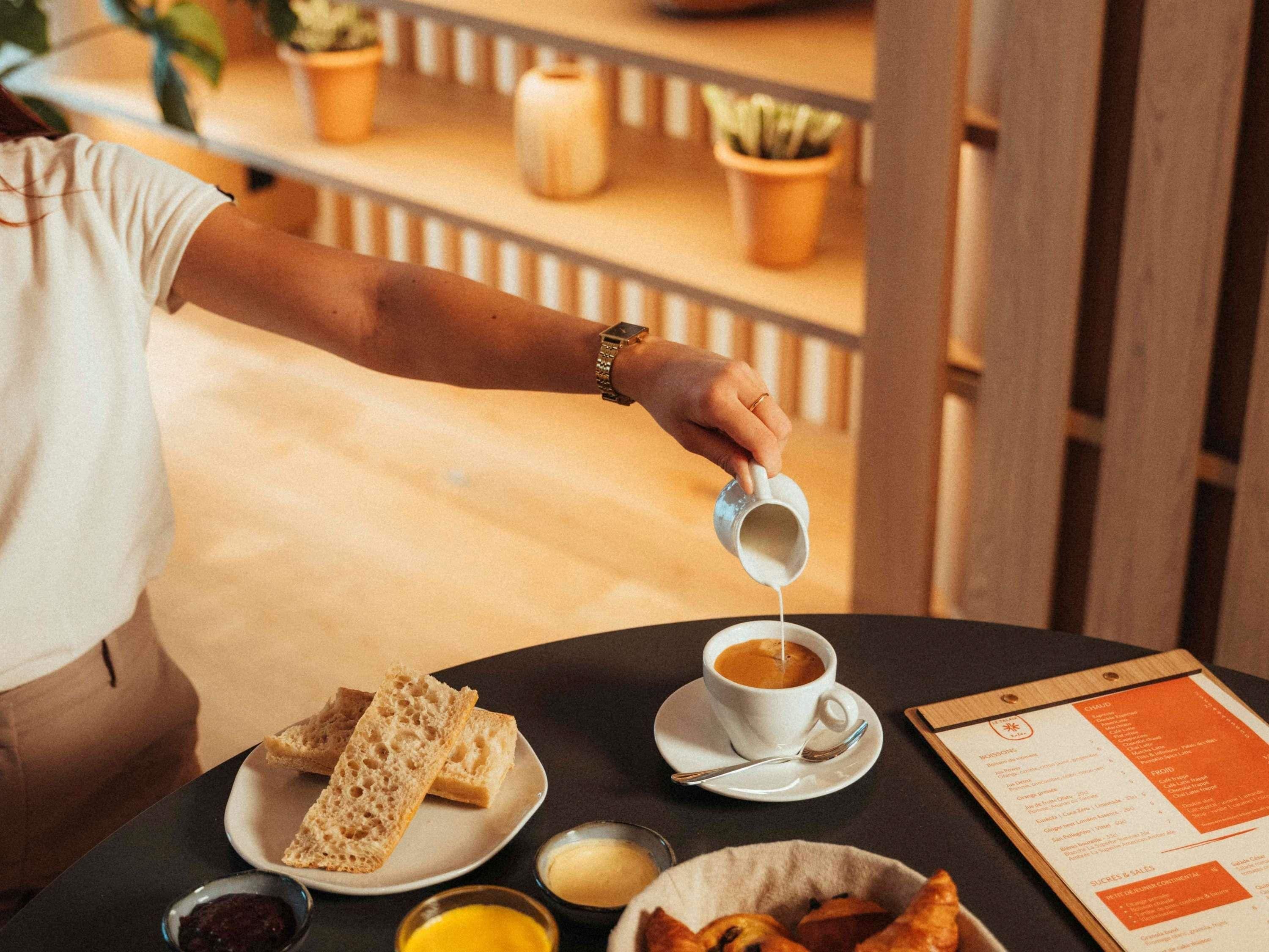A person pouring cream into a cup of coffee on a table with bread and pastries.