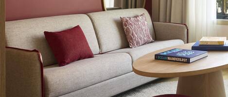 A sofa with two pillows, a round coffee table with a book, and a red ottoman.