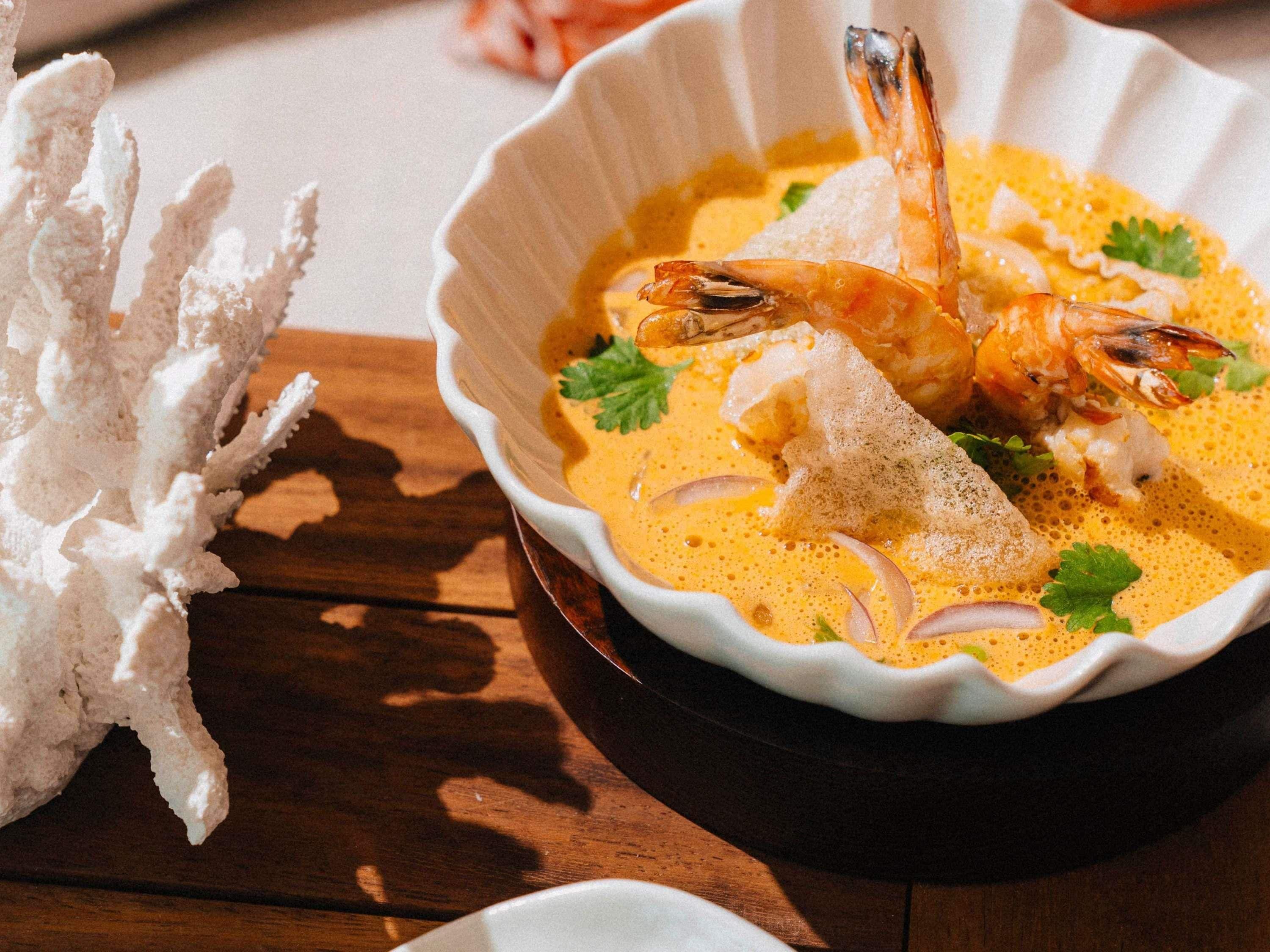 A bowl of soup with shrimp and a white decorative item on a wooden table.