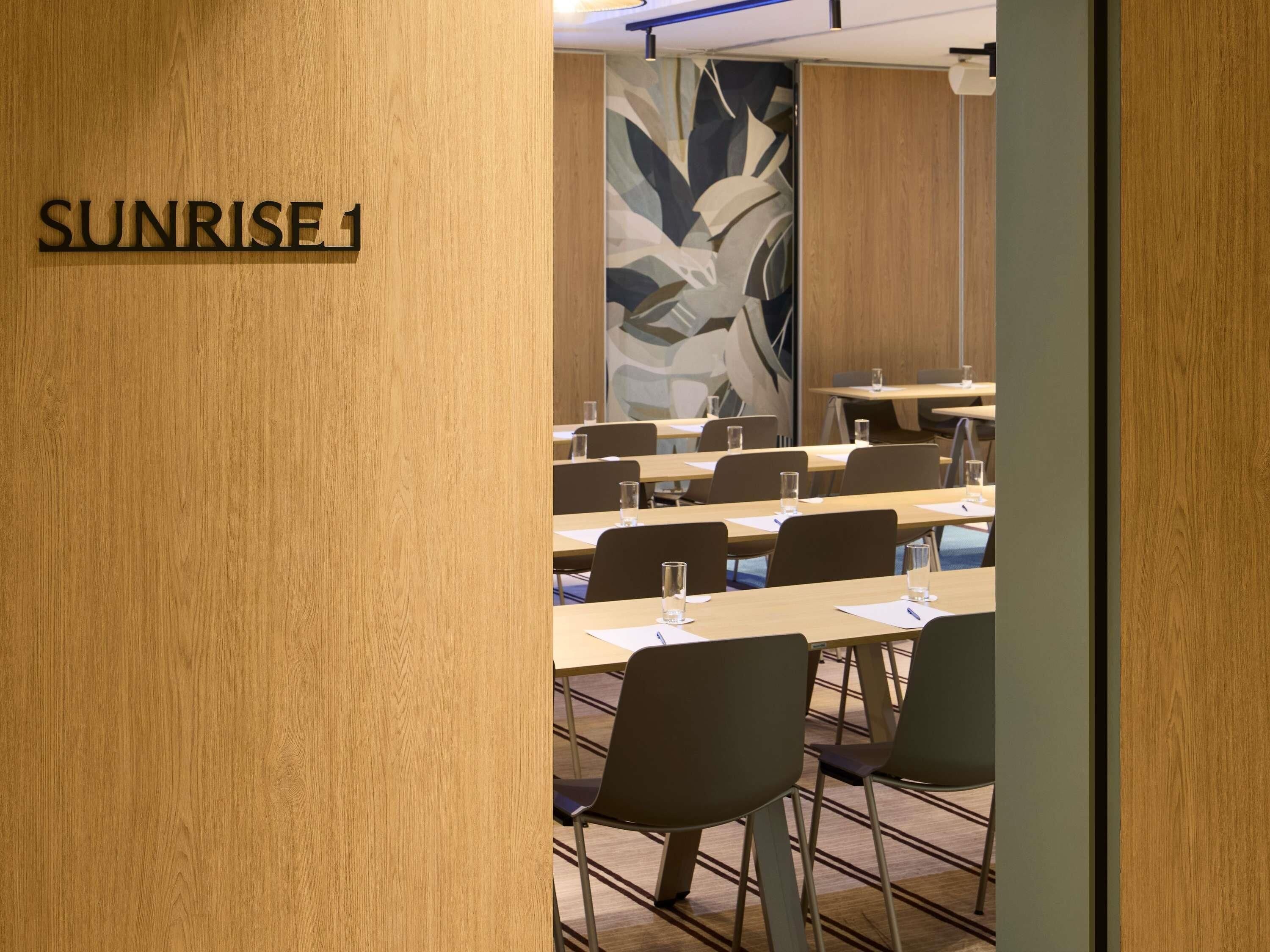Un salón de conferencias con mesas y sillas, una pared de madera con el texto 'SUNRISE 1', y un gran mural abstracto en la pared.