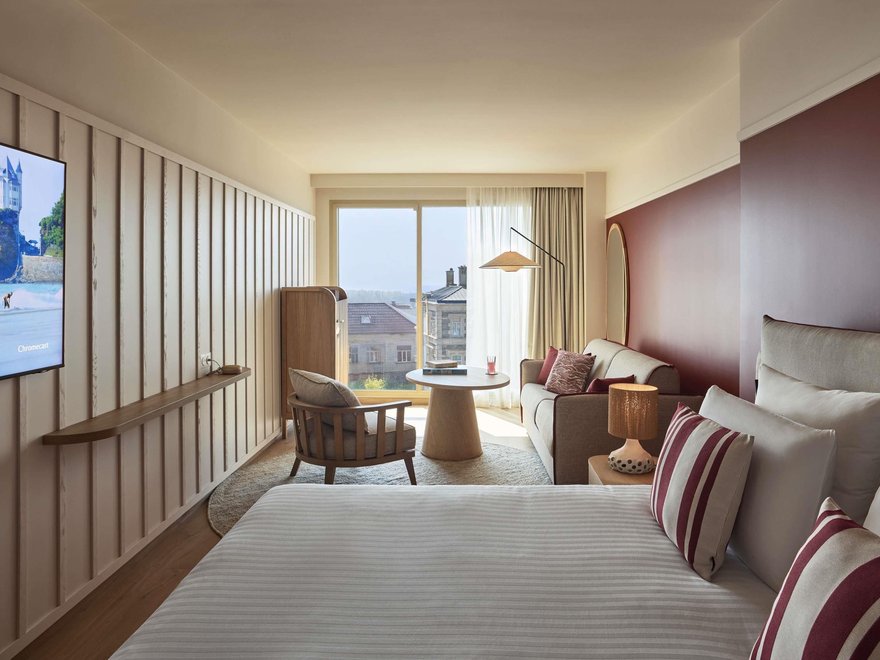 A hotel room with a large bed, a sofa, a chair, a small table, and a TV mounted on the wall.