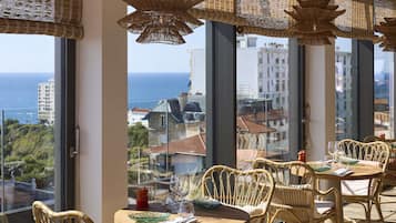 A dining area with wicker furniture, large windows with ocean views, and hanging wicker lamps.