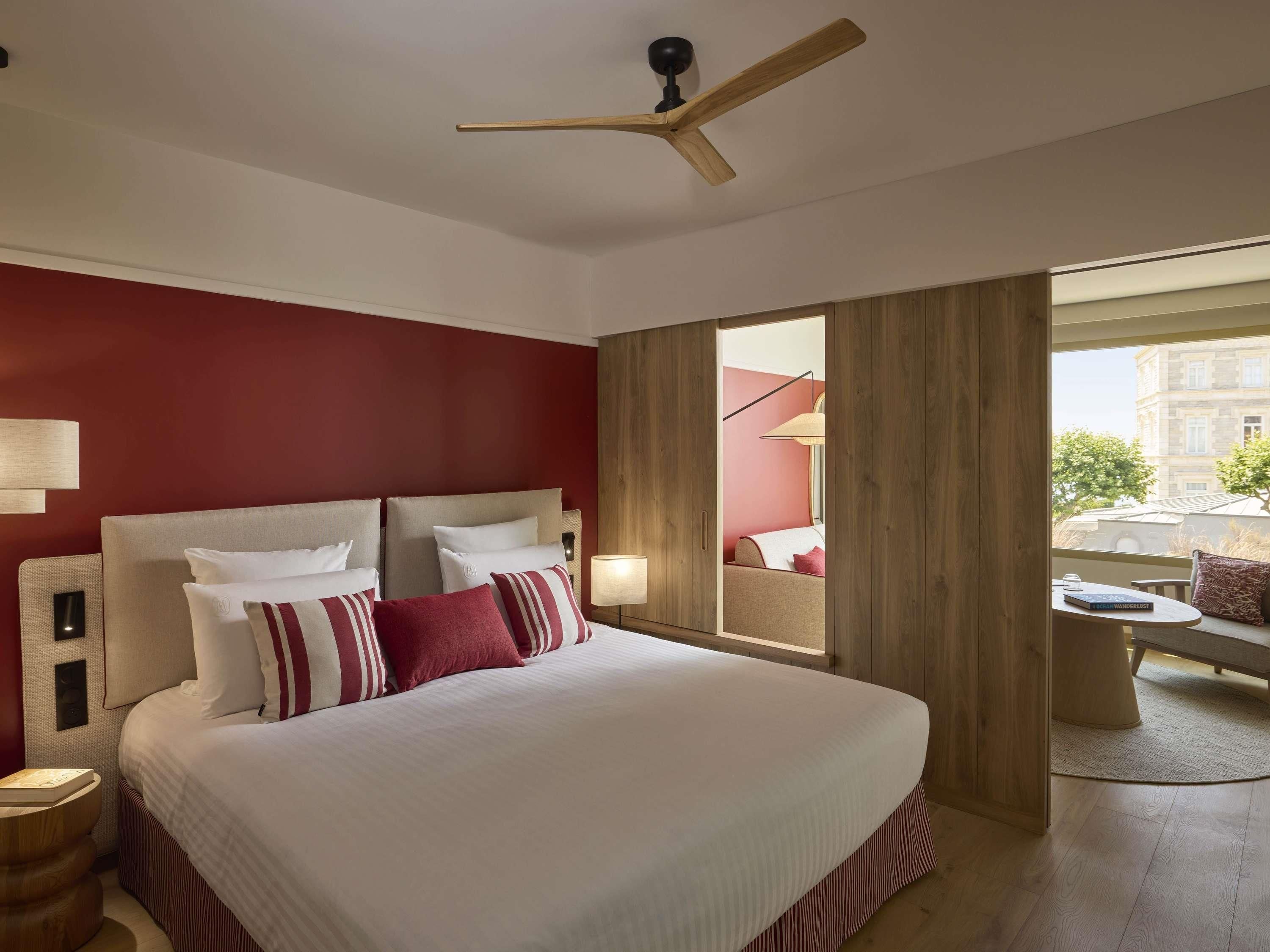 Quarto de hotel com uma cama grande, cabeceira de madeira e ventoinha no teto. Há uma janela com vista para o exterior.