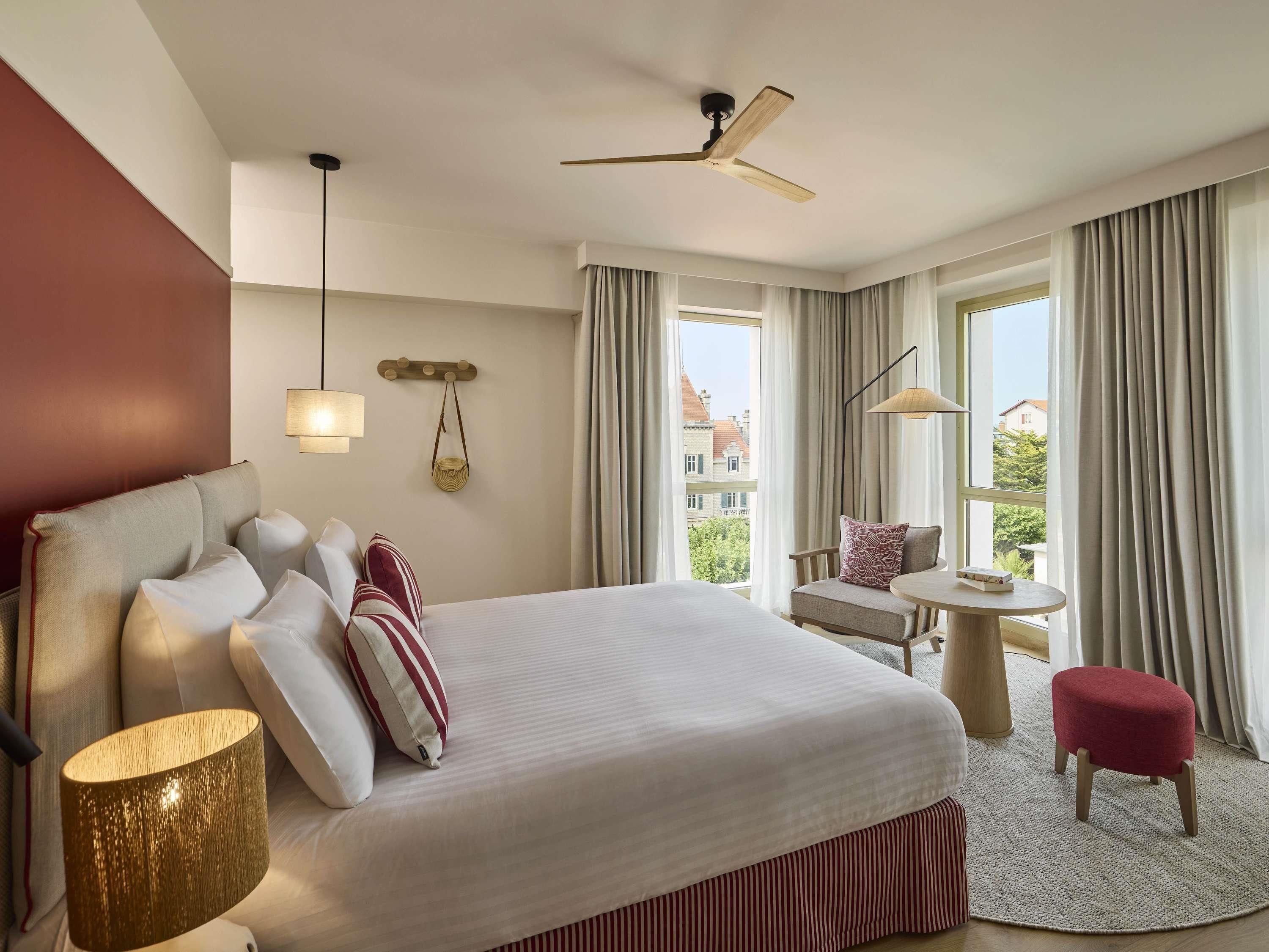 A hotel room with a large bed, a desk, a chair, a small stool, a lamp, a ceiling fan, and a view of buildings through the windows.