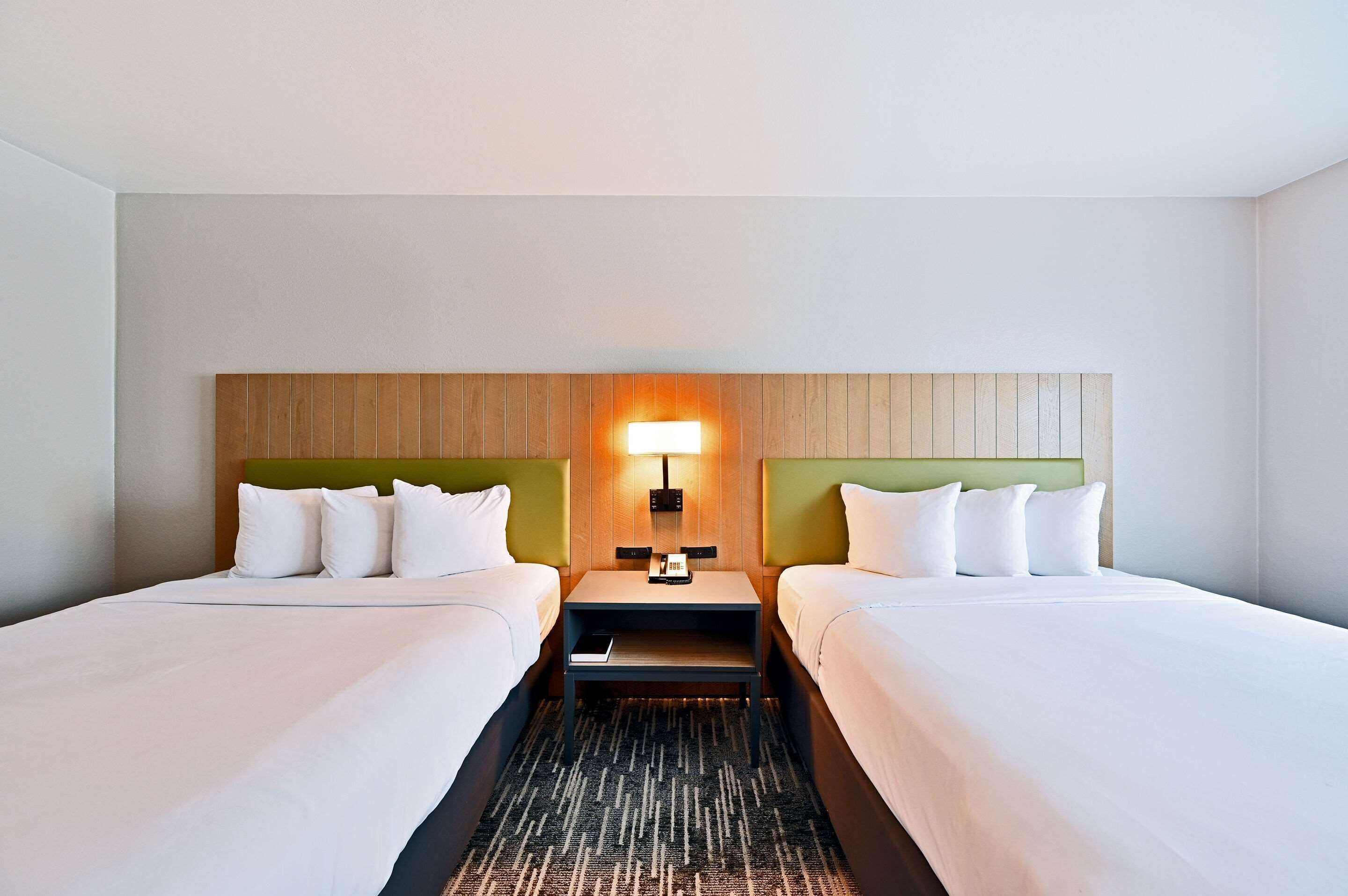 A hotel room with two beds, a nightstand with a lamp, and a headboard with a wooden panel design.