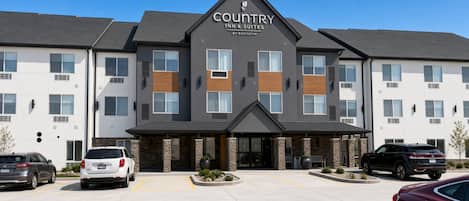 A Country Inn & Suites building with a modern design, featuring a prominent entrance and a parking area with several cars.