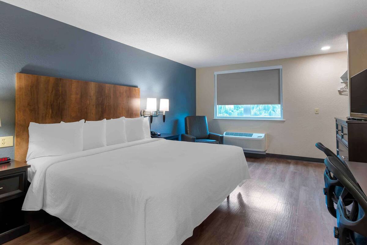 A hotel room with a large bed, a desk with a chair, a TV, and a window with blinds.