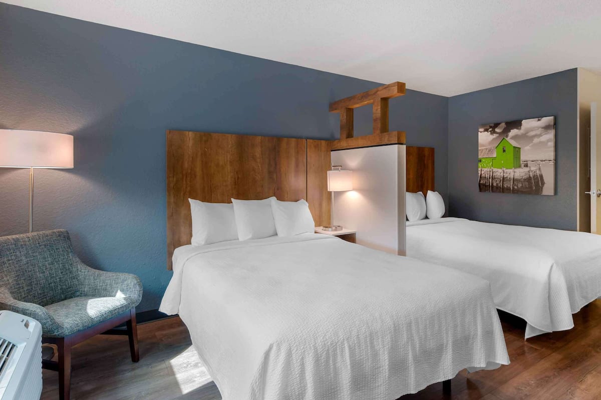 A hotel room with two beds, a wooden headboard, a lamp, a chair, and a painting on the wall.