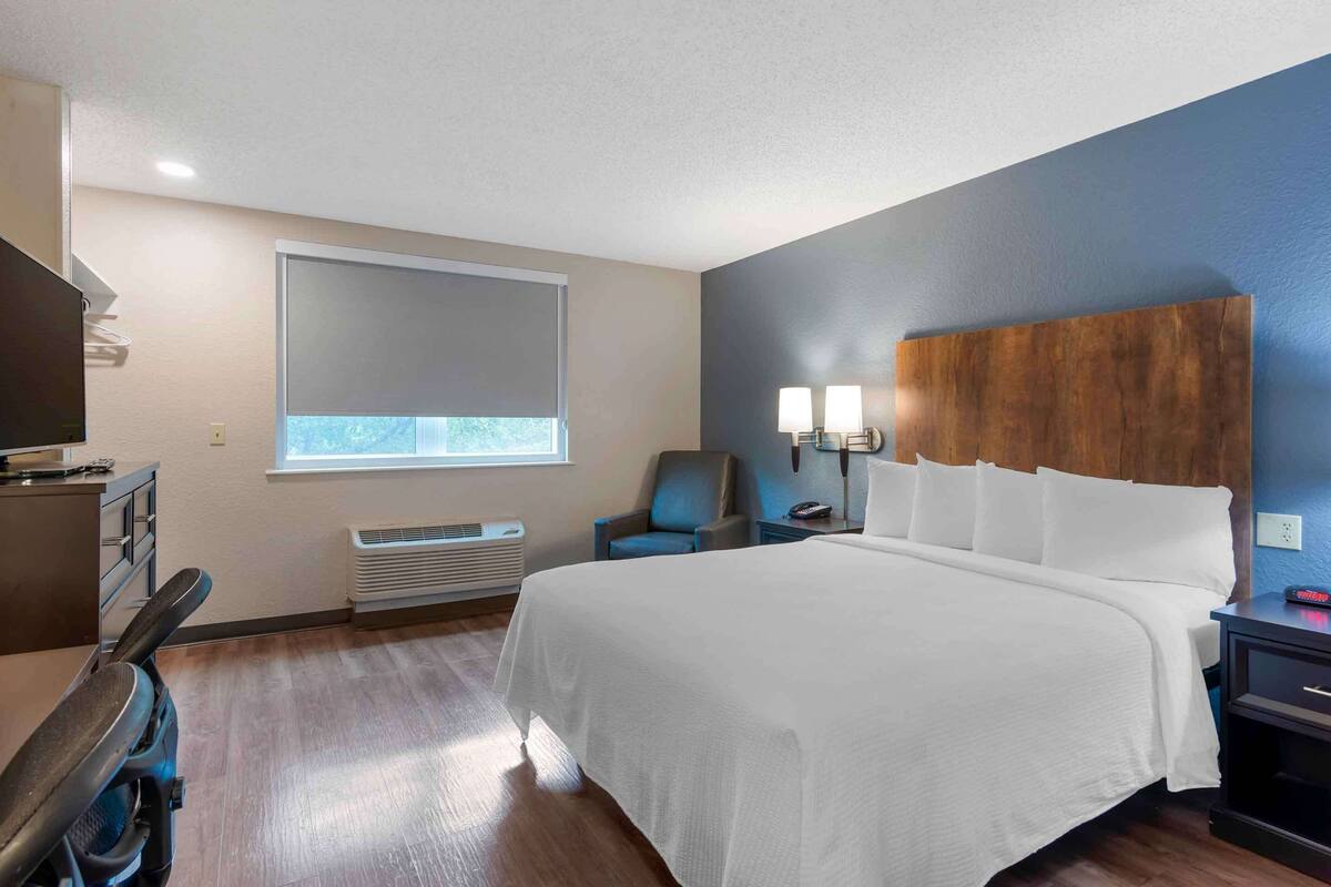 A hotel room with a large bed, a desk with a chair, a TV, a window with blinds, a wall-mounted lamp, and a heater.