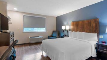 A hotel room with a large bed, a desk with a chair, a TV, a window with blinds, a wall-mounted lamp, and a heater.
