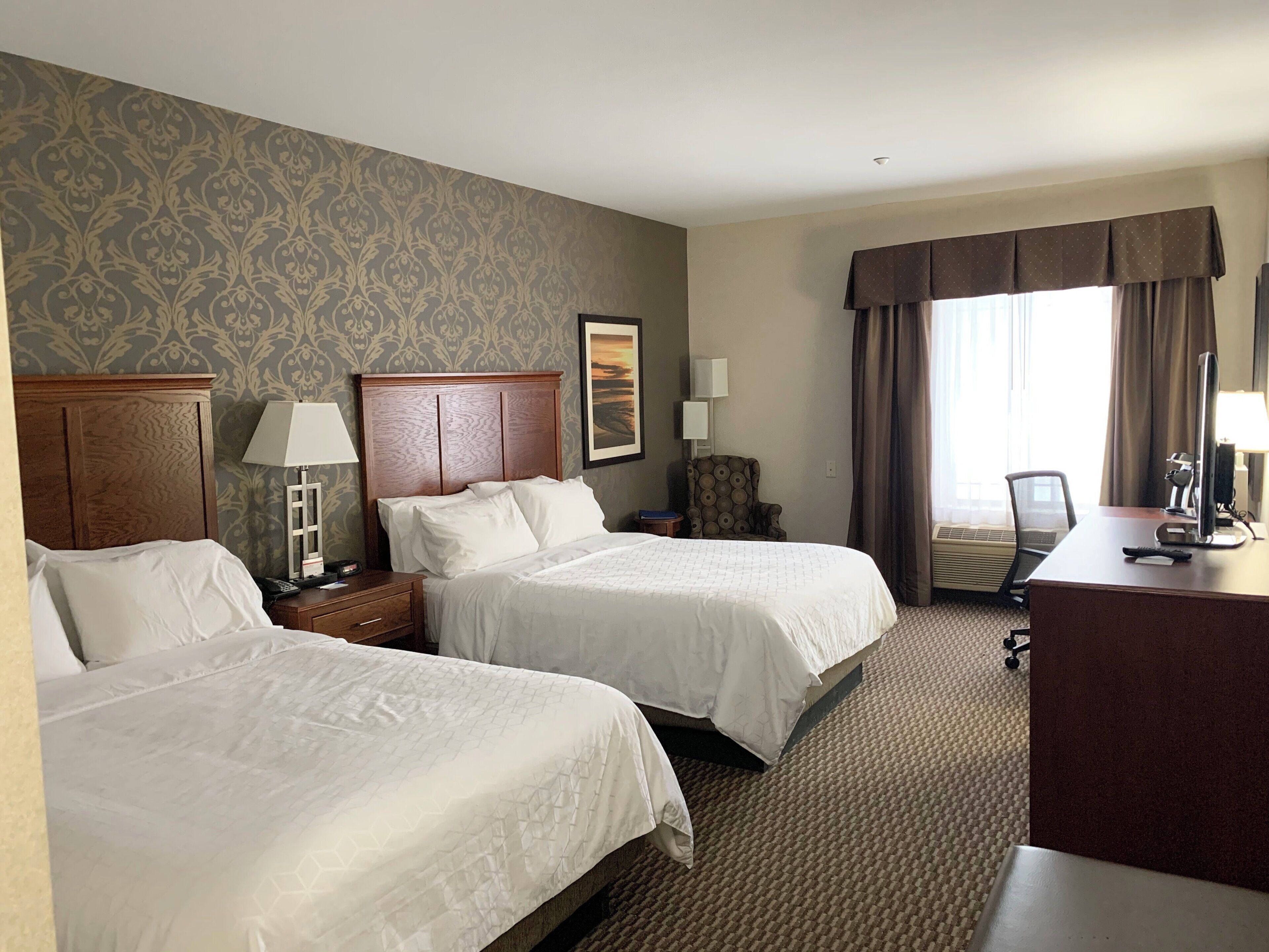 A hotel room with two beds, a desk, a chair, a lamp, a picture on the wall, and a window with curtains.