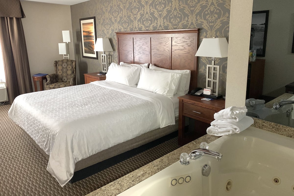 A hotel room with a large bed, a wooden headboard, a nightstand with a lamp, a bathroom with a bathtub, and a mirror.