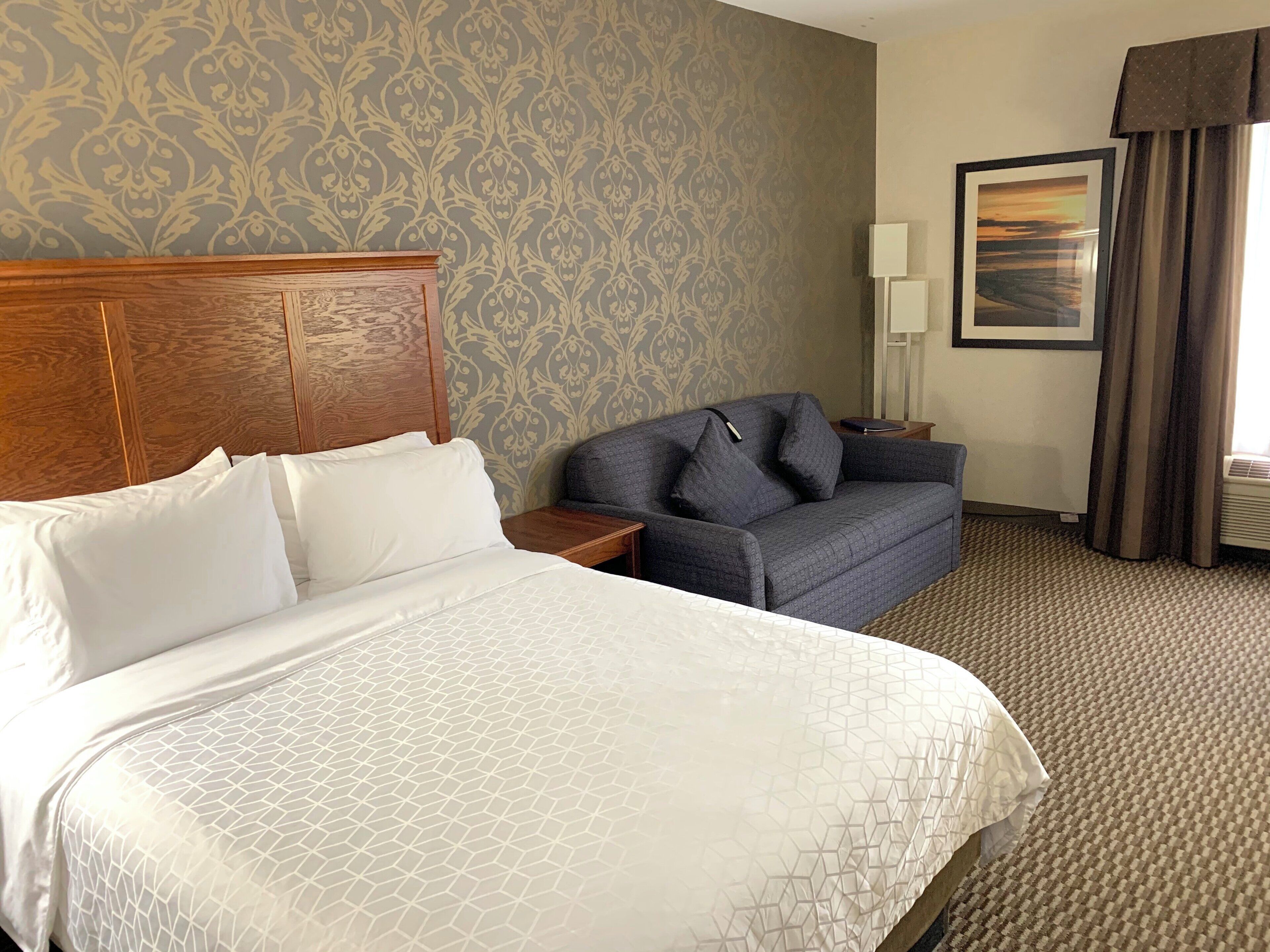 A hotel room with a large bed, a sofa, a nightstand, a lamp, and a framed picture on the wall.