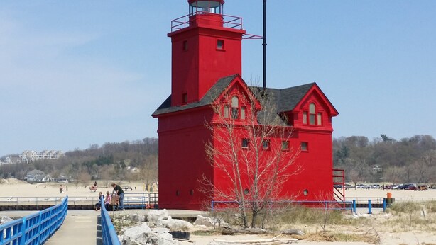 Un phare rouge, doté d’une tour et d’une base, est situé sur une plage de sable, et une promenade bleue mène jusqu’à lui.