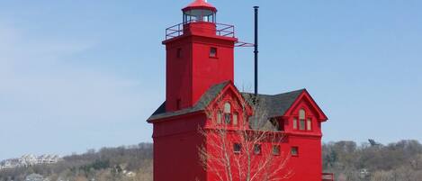 Un faro rojo con una torre y una base, ubicado en una playa arenosa con un paseo marítimo azul que conduce a él.
