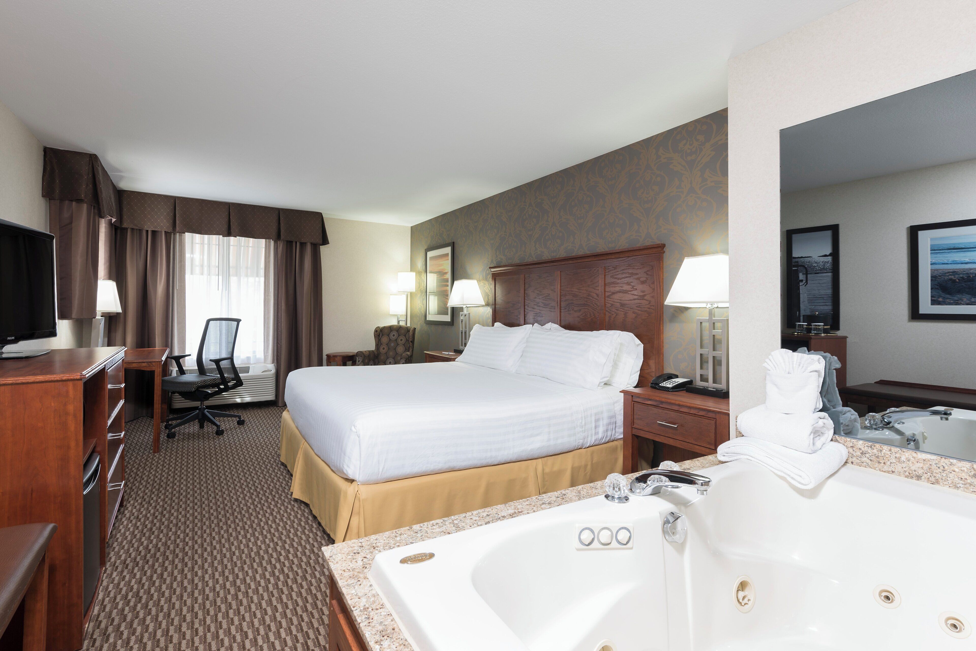 A hotel room with a large bed, a desk with a chair, a television, a bathroom with a bathtub and sink, and a mirror.