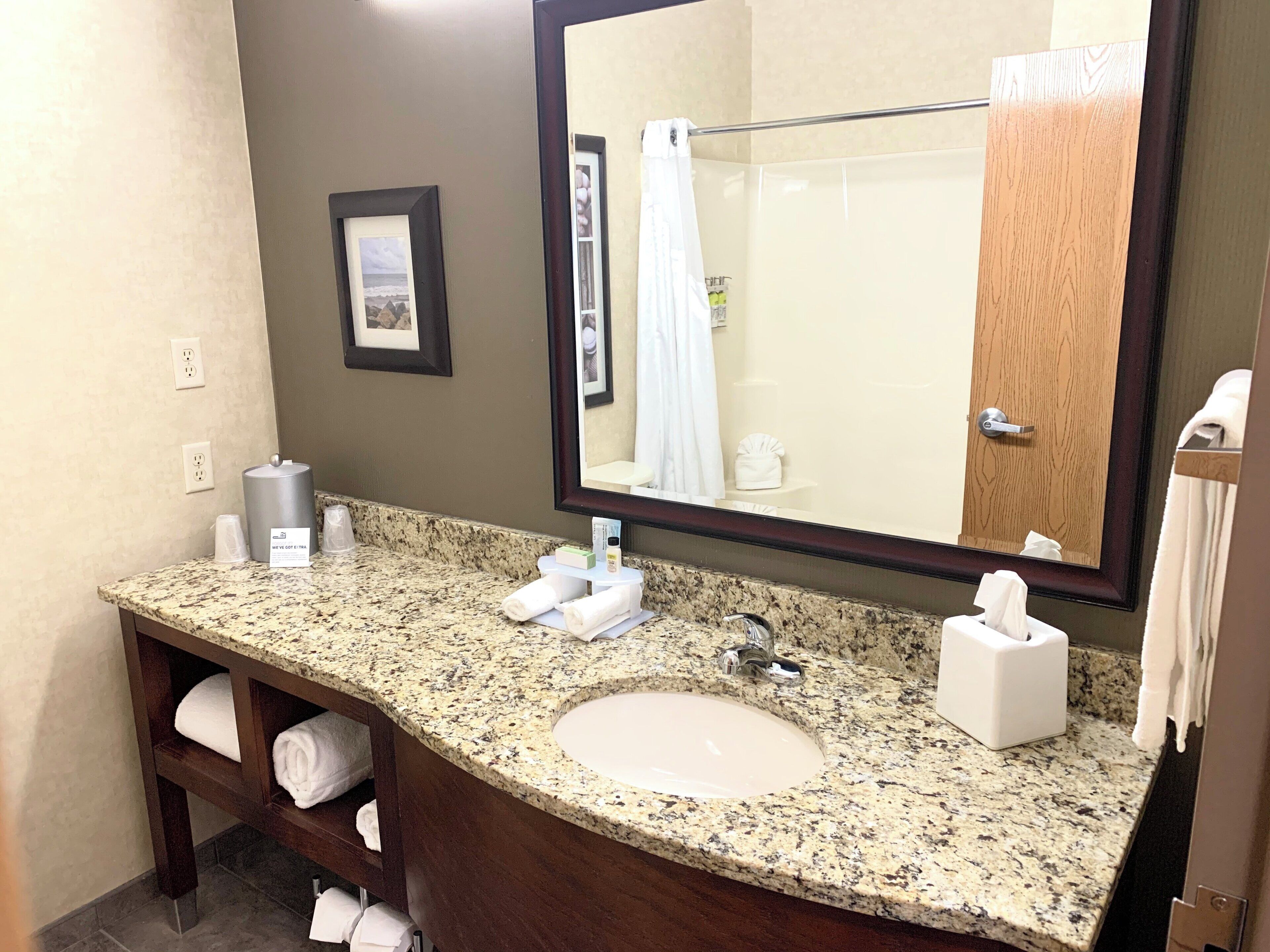 A bathroom with a granite countertop sink, a large mirror, a towel rack, and a shelf with folded towels.