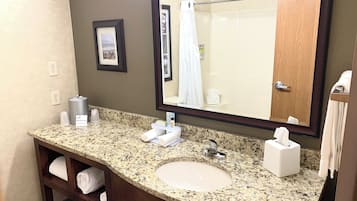 A bathroom with a granite countertop sink, a large mirror, a towel rack, and a shelf with folded towels.