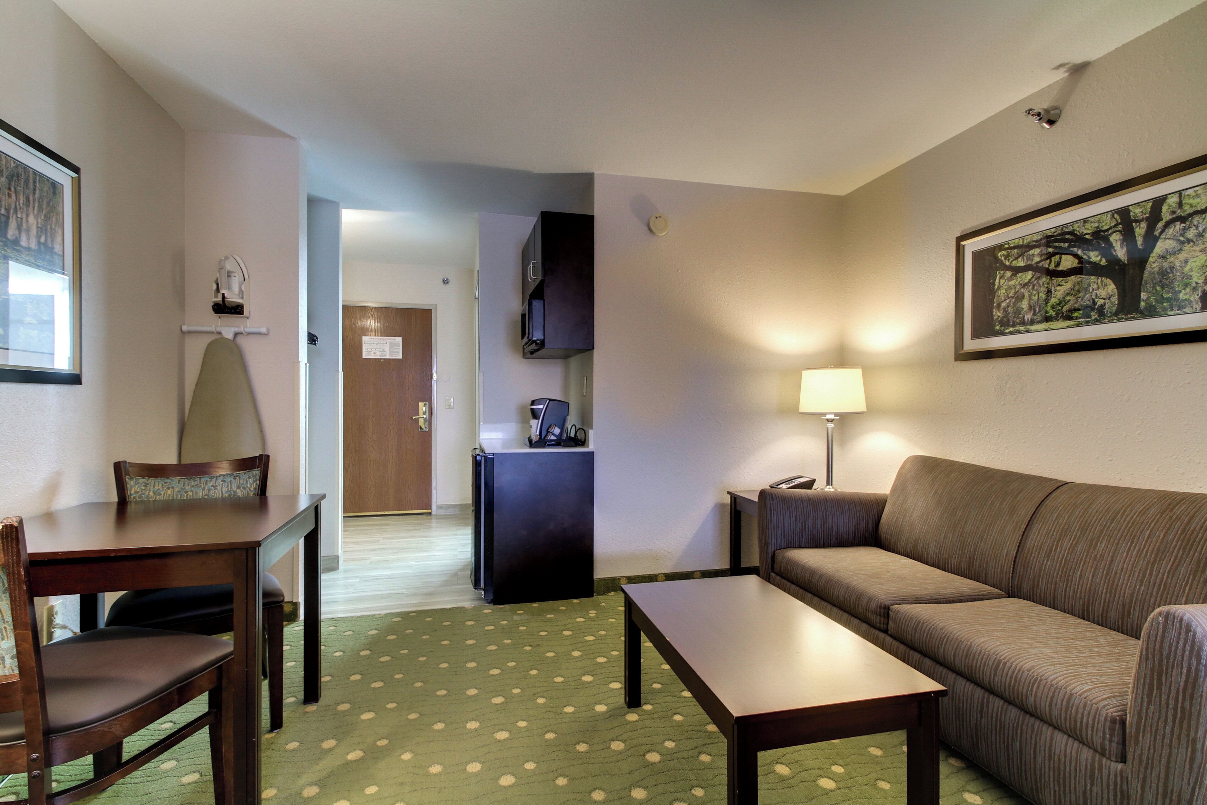 A hotel room with a dining table, a sofa, a small kitchenette, and a door leading to a hallway.