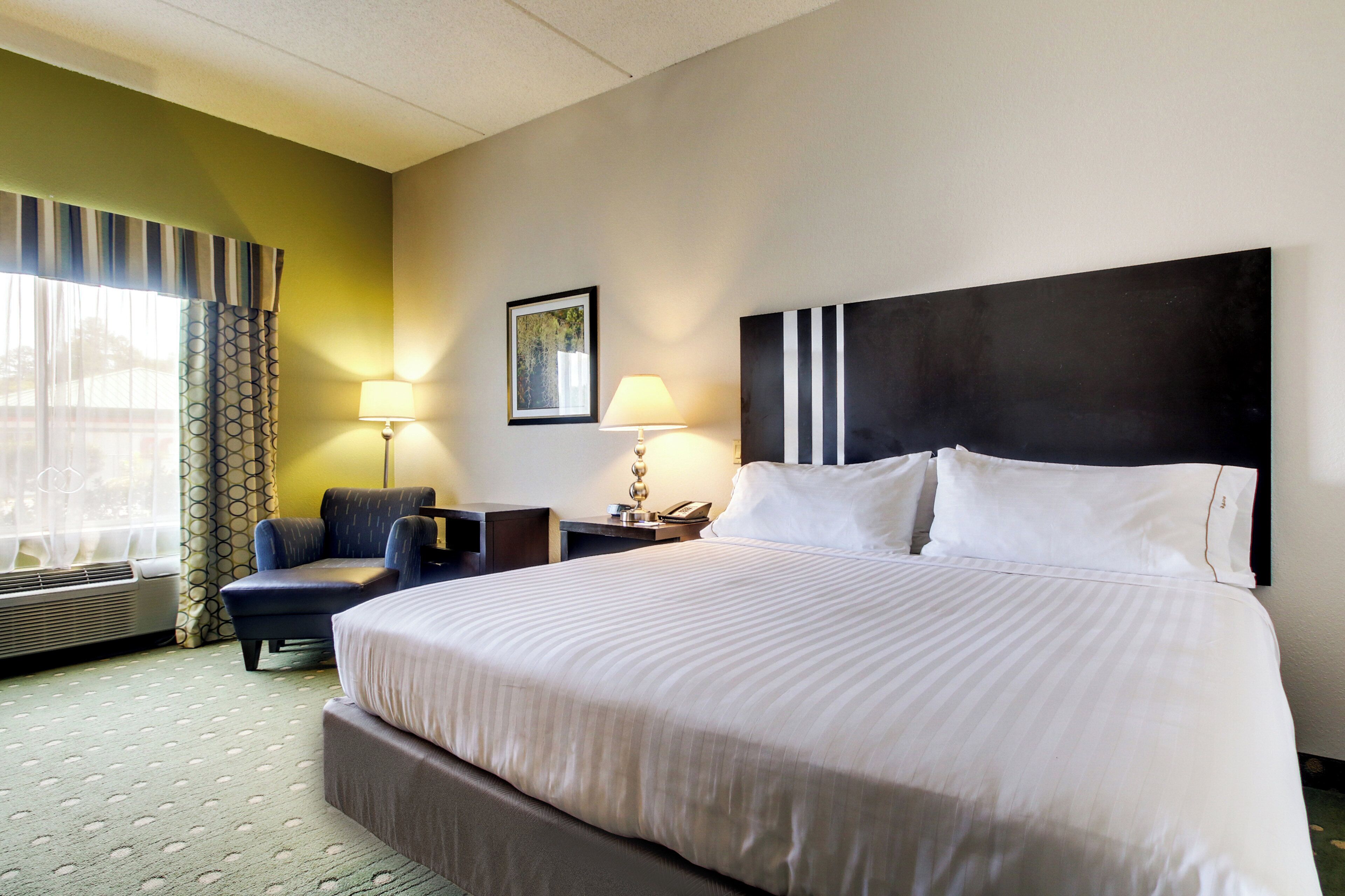 A hotel room with a large bed, a desk, a chair, a window with curtains, a wall-mounted picture, and a bedside lamp.