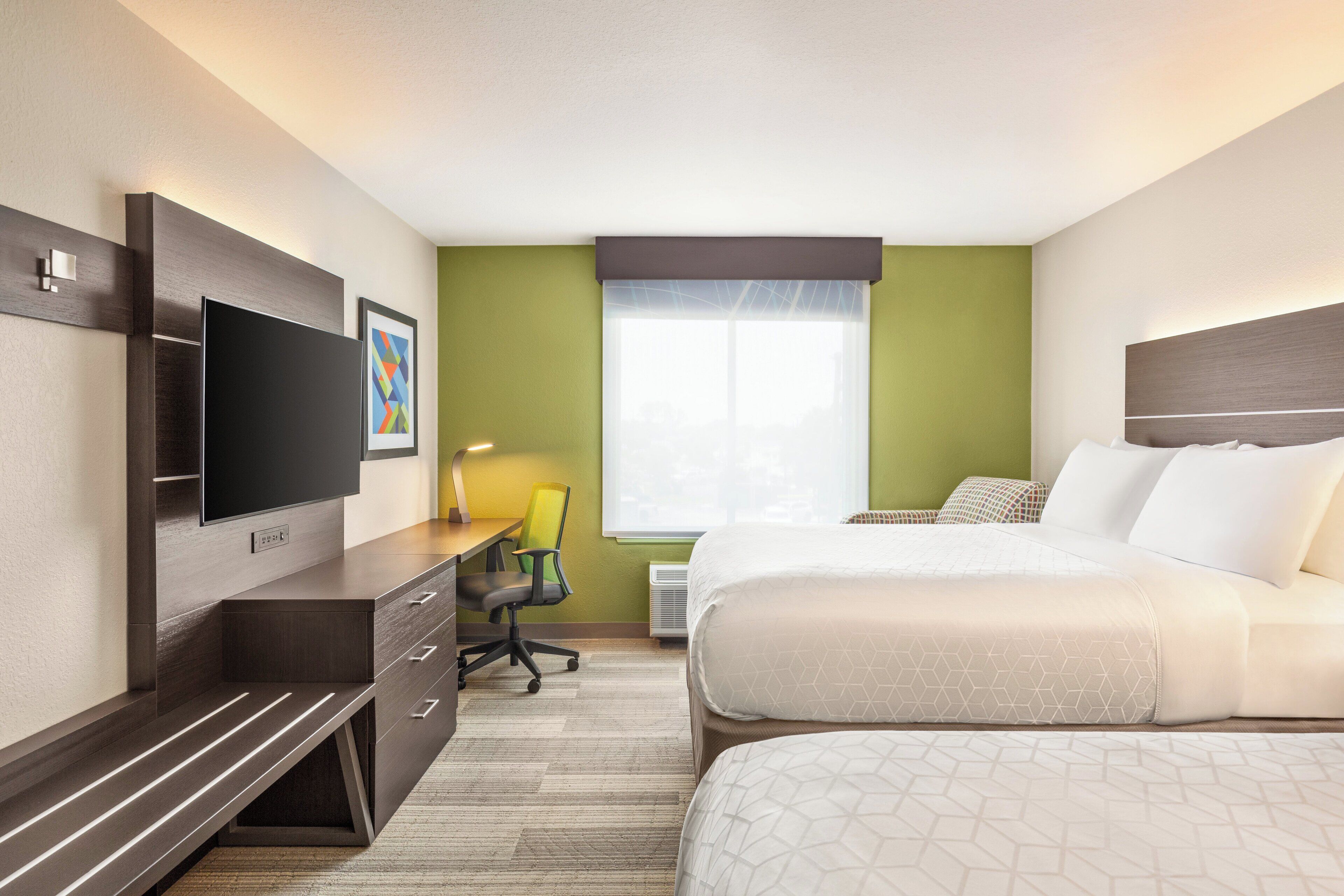 A hotel room with a bed, a desk with a chair, a television, and a window with blinds.