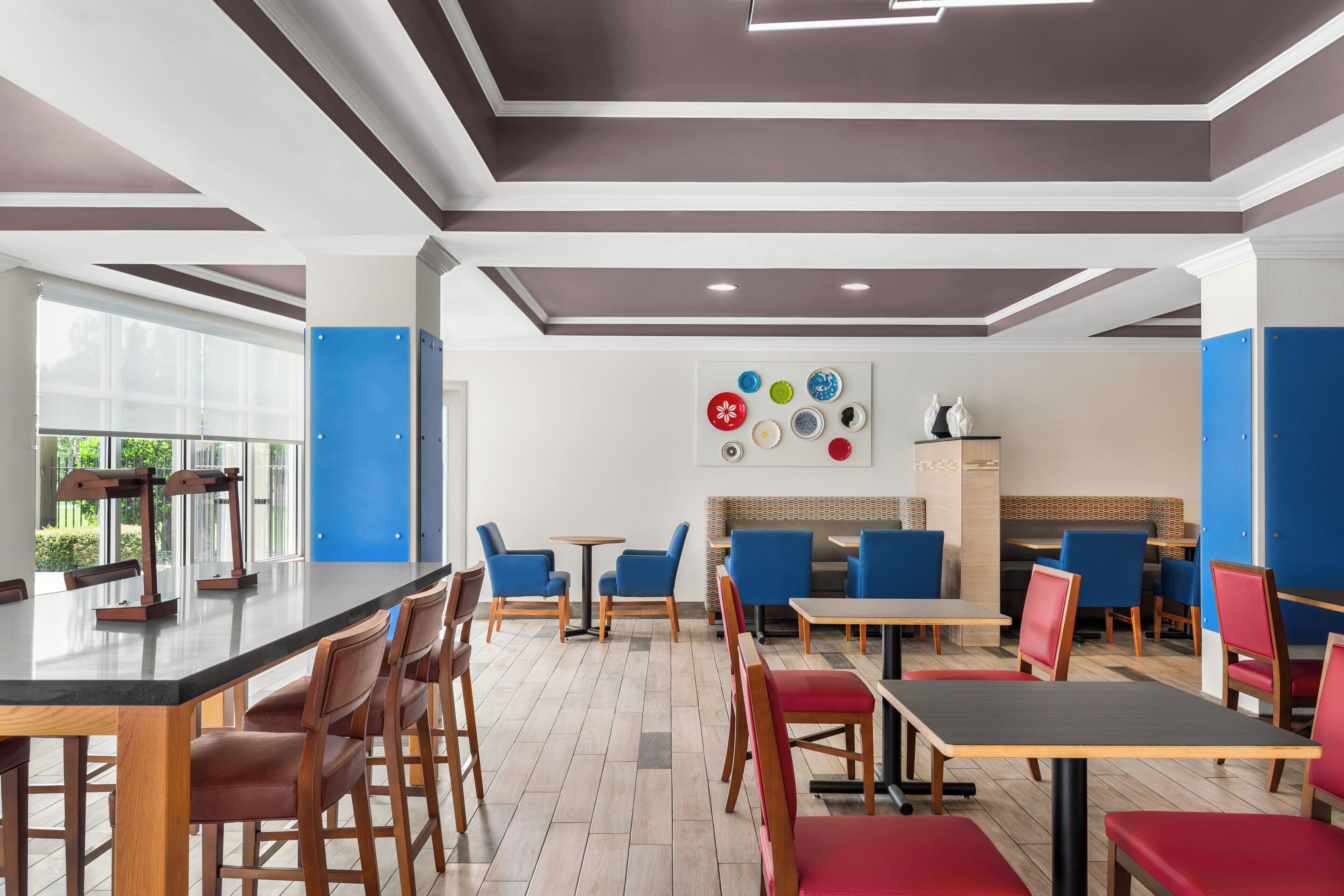 A modern restaurant interior with wooden tables and chairs, a central bar area, and a wall decorated with colorful plates.