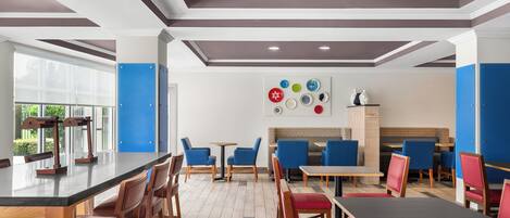 A modern restaurant interior with wooden tables and chairs, a central bar area, and a wall decorated with colorful plates.