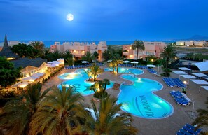 Zona de piscina en un resort, con una luna en el cielo, palmeras y vistas al mar.