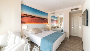 Ett hotellrum med en stor säng, ett skrivbord, en stol, en spegel och en tavla som föreställer ett ökenlandskap.
