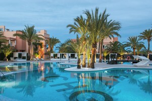 Una piscina con palmeras, sillas de descanso y un edificio al fondo.