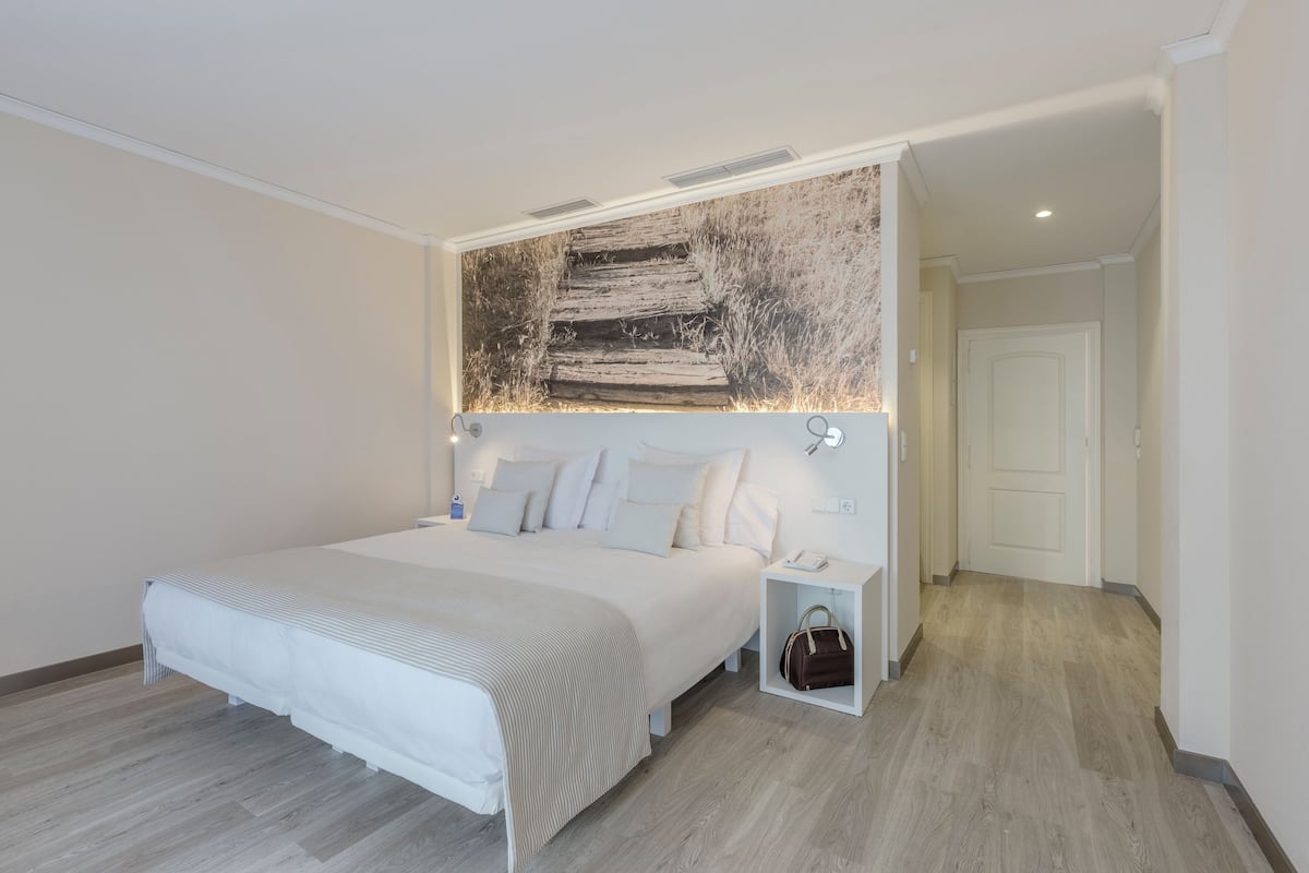 Une chambre moderne avec un grand lit, une tête de lit ornée d’une décoration murale rustique, une table de chevet avec une lampe et une petite boîte de rangement.